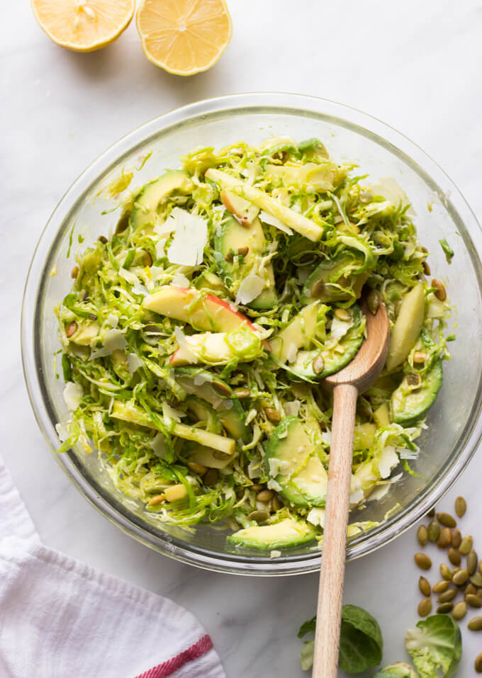 tossed shaved Brussels sprouts with apples, avocado, and dressing in a glass bowl with a wooden spoon.
