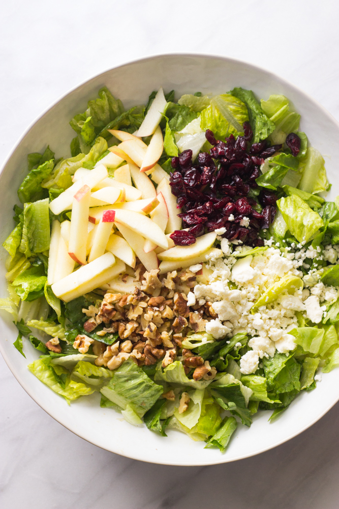 romaine lettuce in a bowl topped with apples, feta, dried cranberries, and walnuts.