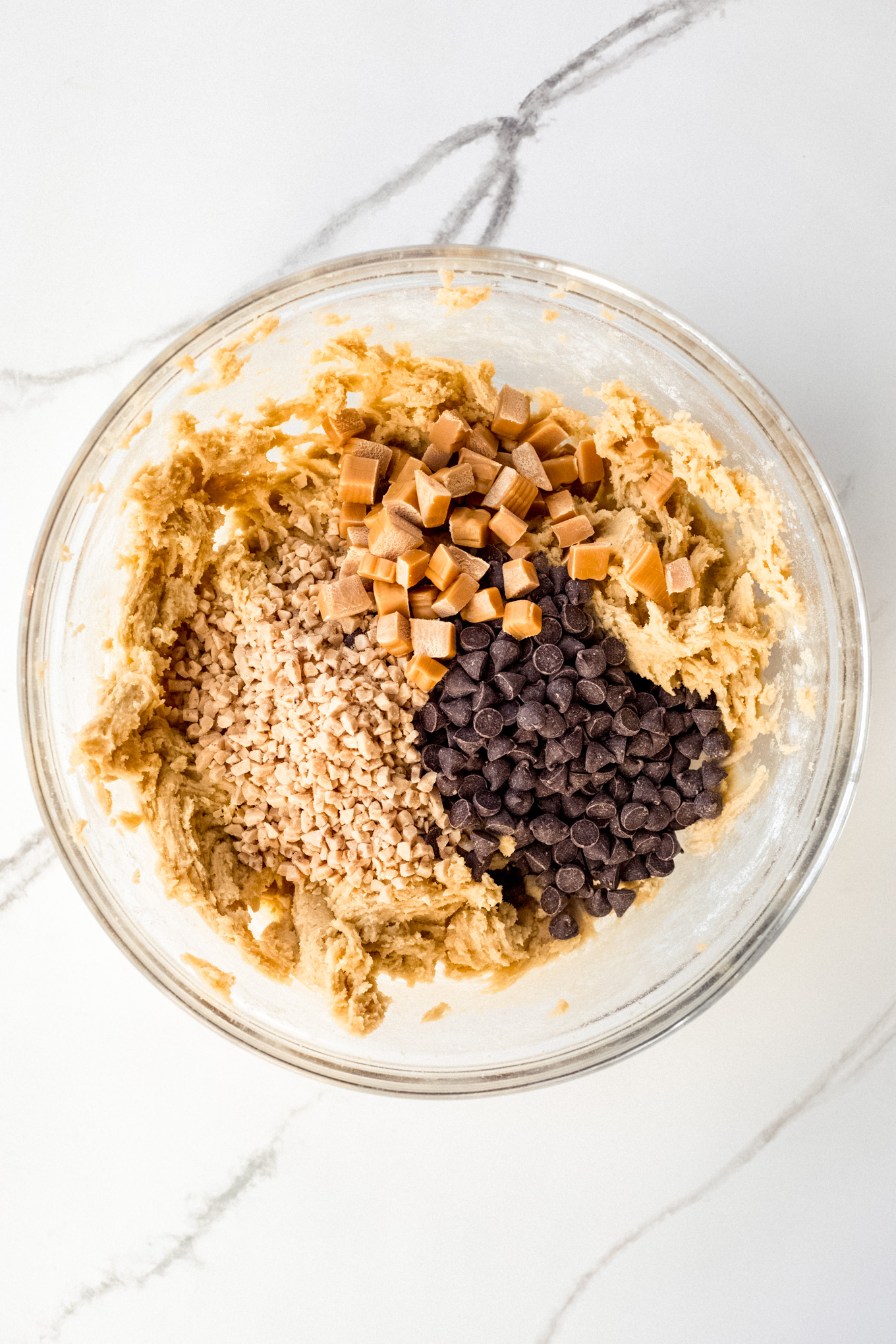 chocolate chips, toffee bits, and caramels over cookies dough in glass bowl.