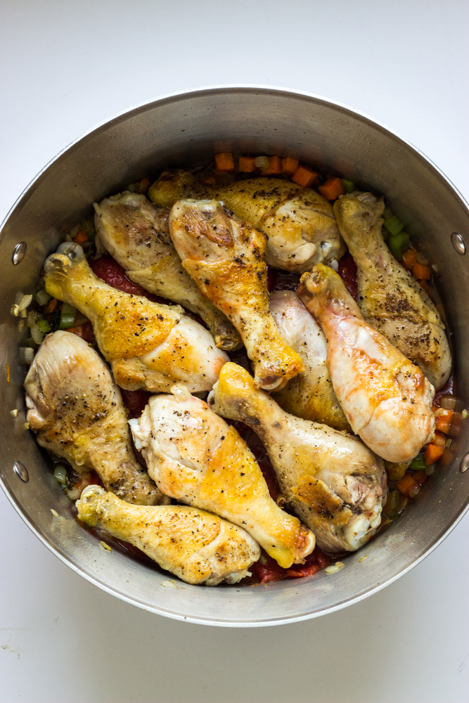 seared chicken drumsticks layered over cooked vegetables inside dutch oven.