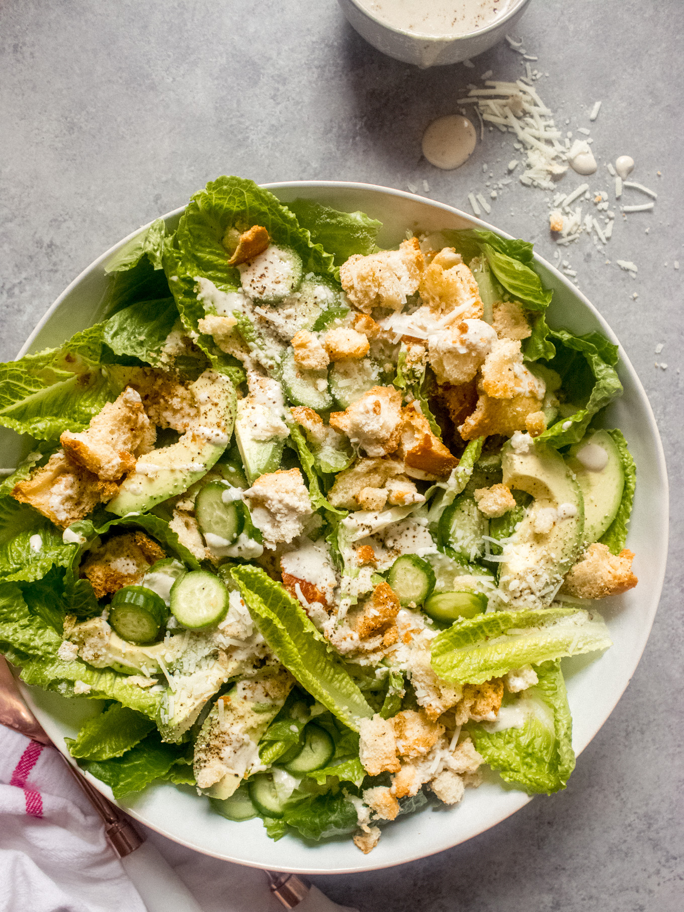 Caesar salad with sliced avocado and cucumbers in a white bowl.