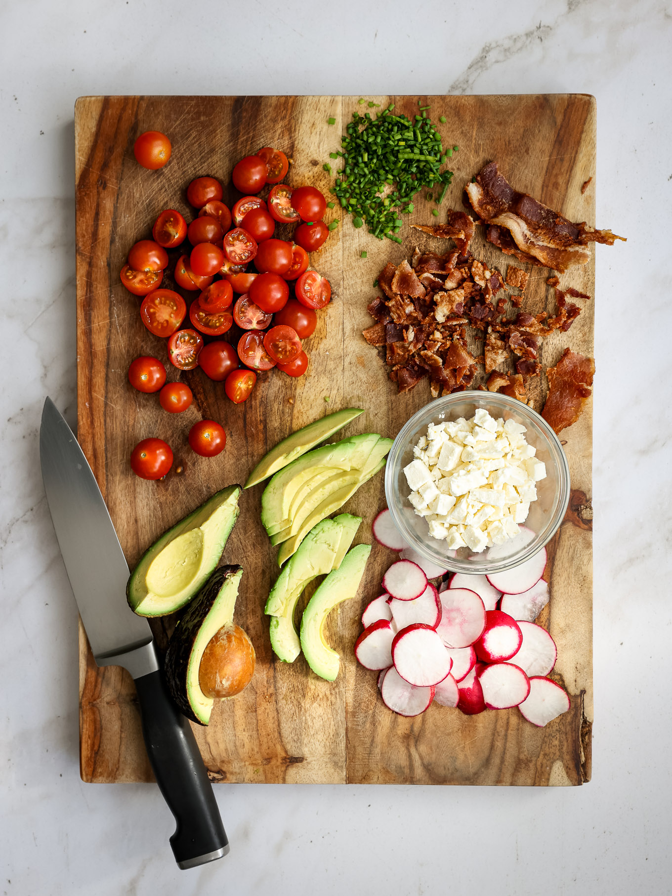 sliced tomatoes, avocado, radishes, bacon, chives, and crumbled feta on a wooden board.