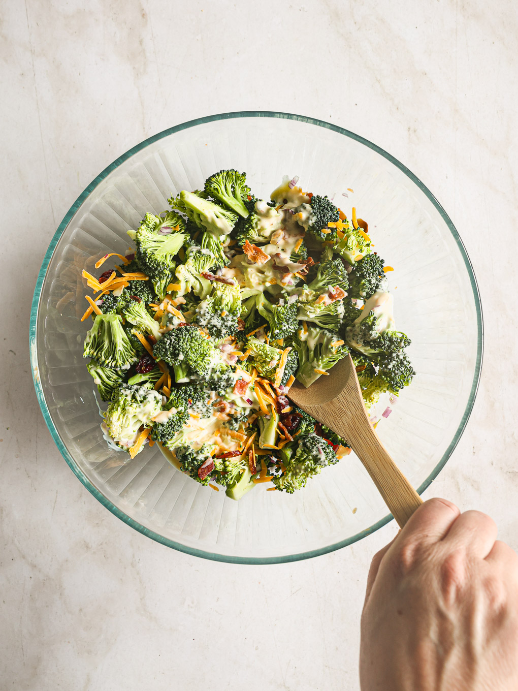 broccoli salad and dressing mixed with wooden spoon in a glass bowl.