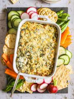 artichoke spinach dip in white baking dish with cut up veggies around and crackers.