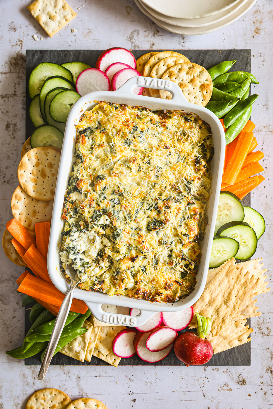 artichoke spinach dip in white baking dish with cut up veggies around and crackers.